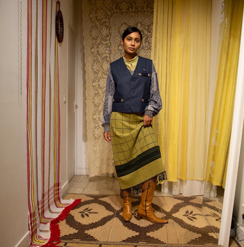 May include: A woman in a navy blue waistcoat with gold buttons, a patterned long-sleeved top, a green plaid skirt with a fringed hem, and brown knee-high boots. The setting is a room with patterned curtains and a decorative rug.