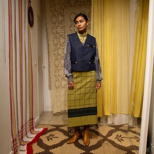 May include: A woman in a navy blue vest, yellow turtleneck, and a long green plaid skirt. The outfit includes a grey sweater with patterned sleeves and brown boots. The background features patterned curtains and a rug.