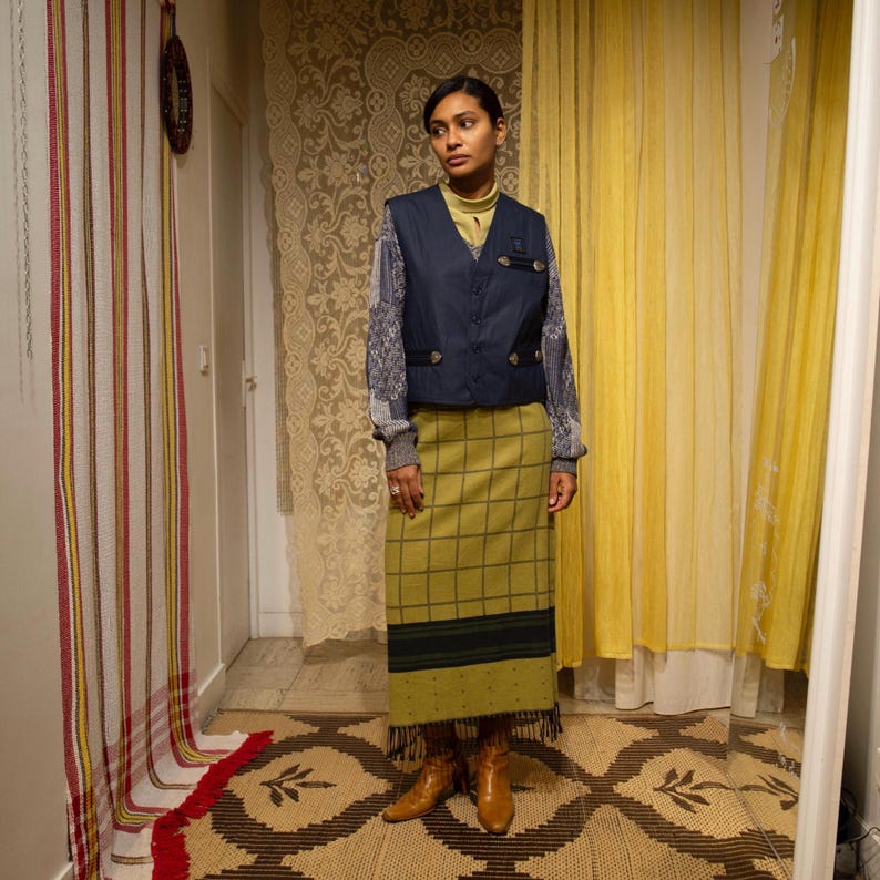 May include: A woman in a navy blue vest, yellow turtleneck, and a long green plaid skirt. The outfit includes a grey sweater with patterned sleeves and brown boots. The background features patterned curtains and a rug.