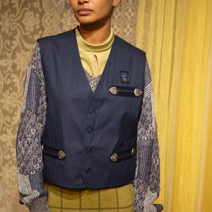 May include: A navy blue waistcoat with silver-coloured details and a small patch. The waistcoat is buttoned and worn over a patterned jumper and a yellow-green top. The outfit includes a plaid skirt.