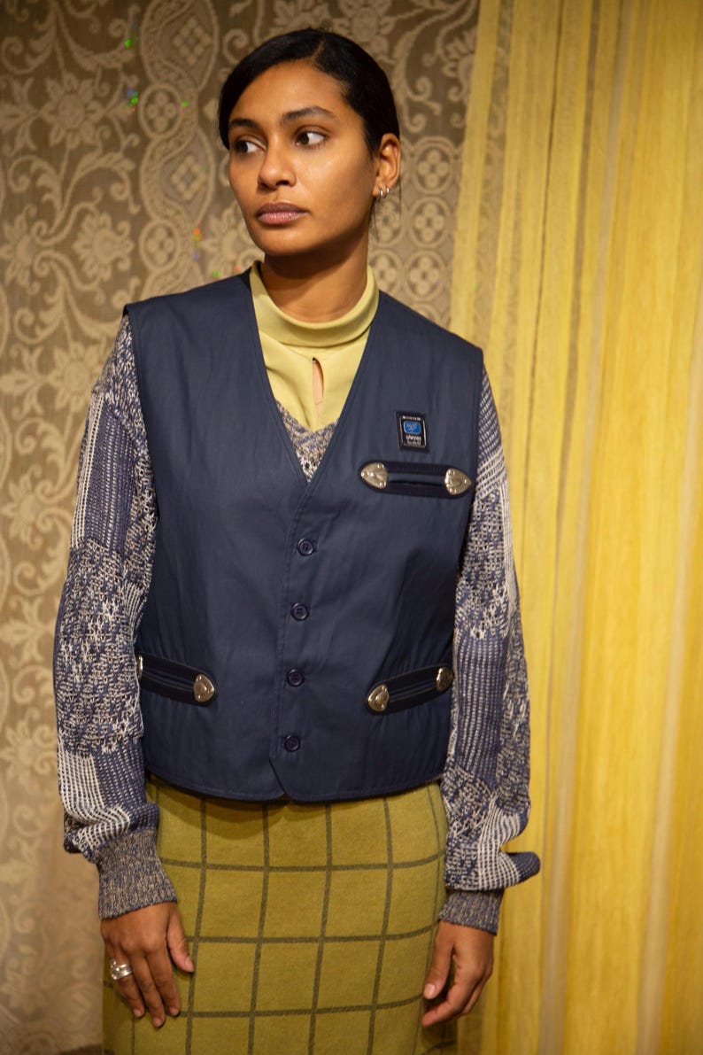 May include: A navy blue waistcoat with silver-coloured details and a small patch. The waistcoat is buttoned and worn over a patterned jumper and a yellow-green top. The outfit includes a plaid skirt.