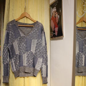 Könnte beinhalten: Ein Vintage-Pullover mit V-Ausschnitt und Patchwork-Design in Blau-, Grau- und Weißtönen hängt an einem Holzbügel. Der Pullover hat gerippte Bündchen und einen grauen Saum. Der Hintergrund umfasst eine Spiegelreflexion und ein Poster.
