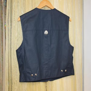 May include: Dark navy vest with a silver emblem on the back. The vest has a classic design with a tailored fit, featuring a button closure and decorative silver buttons. The vest is hanging on a wooden hanger.