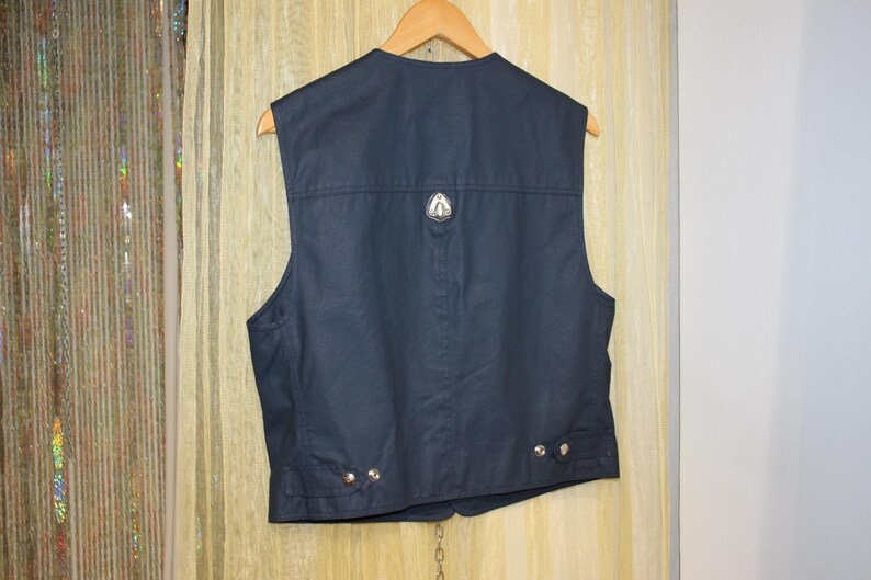 May include: Dark navy vest with a silver emblem on the back. The vest has a classic design with a tailored fit, featuring a button closure and decorative silver buttons. The vest is hanging on a wooden hanger.