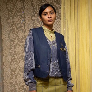 May include: A person wearing a navy blue waistcoat over a patterned jumper and a green plaid skirt. The waistcoat has button closures and metal details. The background features patterned wallpaper and yellow curtains.