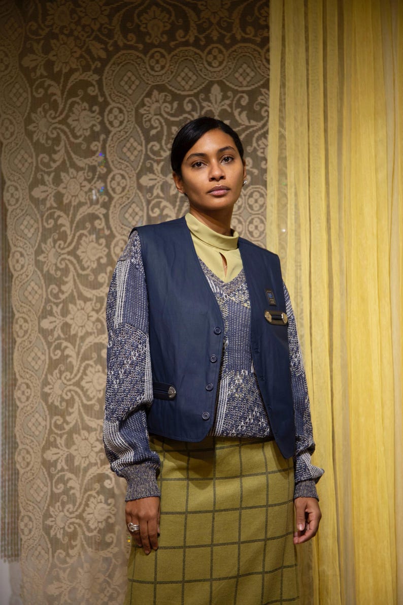 May include: A person wearing a navy blue waistcoat over a patterned jumper and a green plaid skirt. The waistcoat has button closures and metal details. The background features patterned wallpaper and yellow curtains.