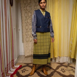 May include: A woman in a navy blue vest over a patterned sweater, a green plaid skirt, and brown boots. The outfit has a vintage style. The background features patterned curtains and a decorative rug.