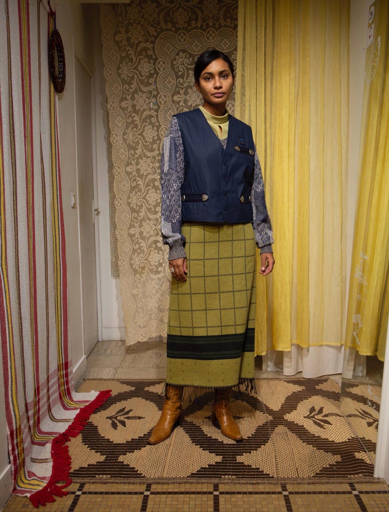 May include: A woman in a navy blue vest over a patterned sweater, a green plaid skirt, and brown boots. The outfit has a vintage style. The background features patterned curtains and a decorative rug.