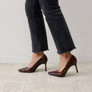 Puede incluir: Un par de zapatos de tac&oacute;n alto con estampado de piel de serpiente en tonos rojos, naranjas y negros. Los zapatos tienen punta puntiaguda y tac&oacute;n de aguja.