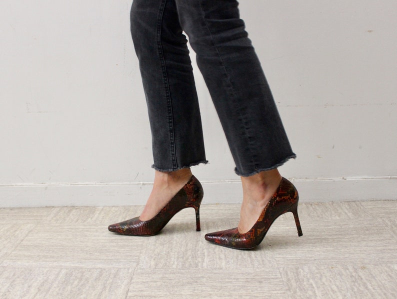 Puede incluir: Un par de zapatos de tac&oacute;n alto con estampado de piel de serpiente en tonos rojos, naranjas y negros. Los zapatos tienen punta puntiaguda y tac&oacute;n de aguja.