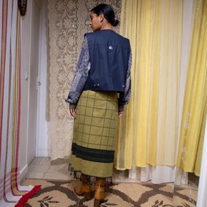 May include: A navy blue vest with silver fastenings is worn over a patterned sweater. An olive green plaid skirt with a black stripe and fringe is paired with brown boots. The outfit is displayed in a room with patterned curtains and a rug.