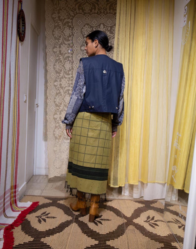May include: A navy blue vest with silver fastenings is worn over a patterned sweater. An olive green plaid skirt with a black stripe and fringe is paired with brown boots. The outfit is displayed in a room with patterned curtains and a rug.