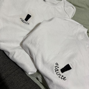 Puede incluir: Dos camisetas blancas con bordados de pintas negras y la palabra "sl&aacute;inte" en cursiva. Las camisetas tienen cuello redondo y una etiqueta que dice "BEWITCH". Las camisetas est&aacute;n dobladas sobre una superficie verde claro.