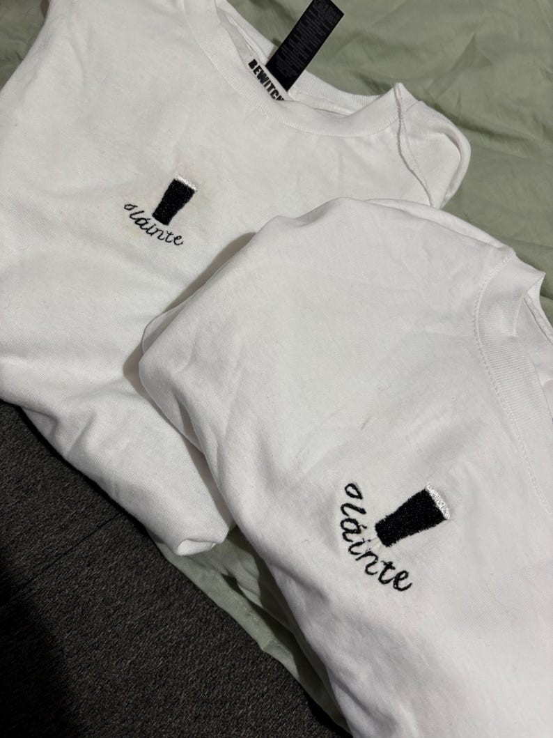 Puede incluir: Dos camisetas blancas con bordados de pintas negras y la palabra "sl&aacute;inte" en cursiva. Las camisetas tienen cuello redondo y una etiqueta que dice "BEWITCH". Las camisetas est&aacute;n dobladas sobre una superficie verde claro.