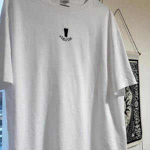 Puede incluir: Camiseta blanca con un dise&ntilde;o bordado en negro de una pinta de cerveza y la palabra "olainte" debajo. La camiseta est&aacute; colgada en una percha negra.