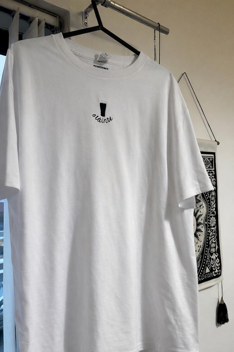 Puede incluir: Camiseta blanca con un dise&ntilde;o bordado en negro de una pinta de cerveza y la palabra "olainte" debajo. La camiseta est&aacute; colgada en una percha negra.