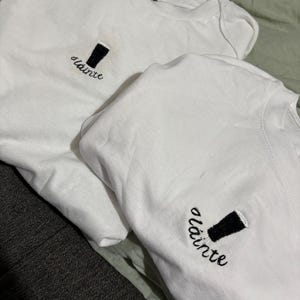 Puede incluir: Dos camisetas blancas con un vaso de pinta negro bordado y la palabra "sl&aacute;inte" en cursiva. Una etiqueta negra que dice "BEWITCH" es visible en una camiseta.