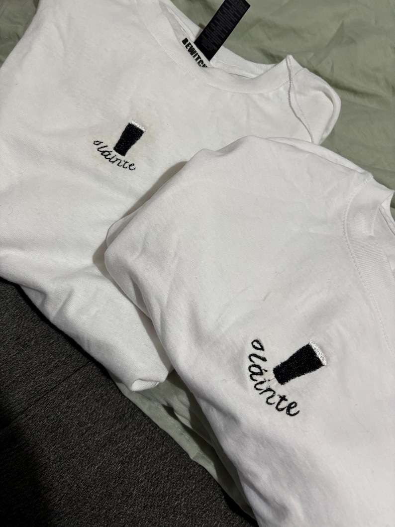 Puede incluir: Dos camisetas blancas con un vaso de pinta negro bordado y la palabra "sl&aacute;inte" en cursiva. Una etiqueta negra que dice "BEWITCH" es visible en una camiseta.