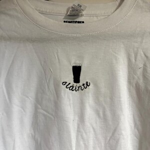 Puede incluir: Camiseta blanca con una pinta de cerveza bordada en negro y la palabra "Sl&aacute;inte" en cursiva. La camiseta tiene cuello redondo y mangas cortas. La marca "REWITCHED" est&aacute; impresa en la etiqueta.