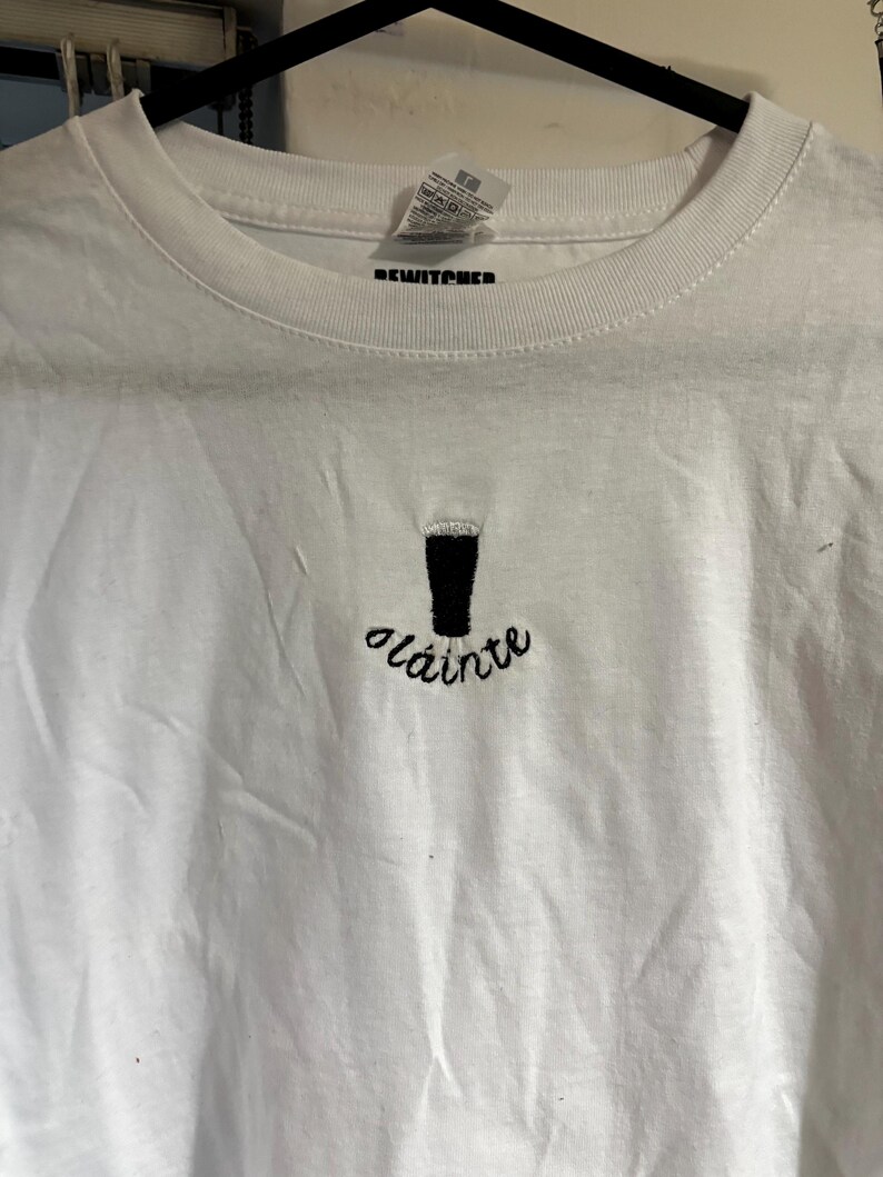 Puede incluir: Camiseta blanca con una pinta de cerveza bordada en negro y la palabra "Sl&aacute;inte" en cursiva. La camiseta tiene cuello redondo y mangas cortas. La marca "REWITCHED" est&aacute; impresa en la etiqueta.