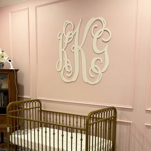 May include: A gold metal crib with a white mattress in a nursery with pink walls and white trim. A white wooden monogram with the letters "K.G." is on the wall above the crib.