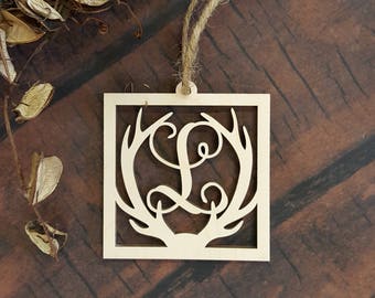 May include: A wooden ornament with a square shape and a cutout design of antlers and the letter L. The ornament is hanging from a twine string.