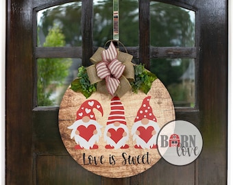 May include: A round wooden door hanger featuring three gnomes holding red hearts. The gnomes are wearing red and white hats with white beards. The door hanger has the text "Love is Sweet" and a small logo that says "Born Love".