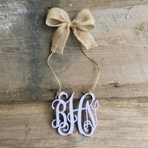 May include: A light purple wooden monogram with the letters "BHA" in a cursive font. The monogram is hanging from a twine string with a burlap bow at the top.