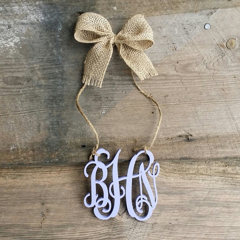 May include: A light purple wooden monogram with the letters "BHA" in a cursive font. The monogram is hanging from a twine string with a burlap bow at the top.