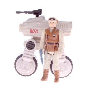 May include: A vintage Star Wars action figure of a soldier in a white and brown uniform, standing next to a white and gray walker vehicle with a black cannon. The figure is holding a blue weapon.
