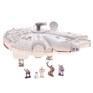 Puede incluir: Una maqueta blanca de la nave espacial Halcón Milenario con una torreta de cañón gris y una antena de radar. Varias figuras en miniatura de personajes están colocadas delante de la nave espacial.