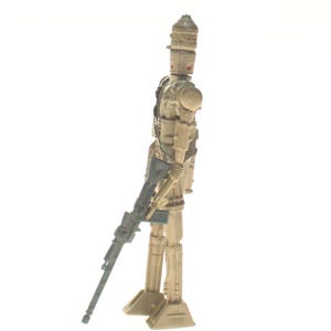 May include: A beige and gray plastic action figure of a Star Wars character, a droid, holding a blue and gray blaster rifle. The droid has a dome-shaped head with a red eye.