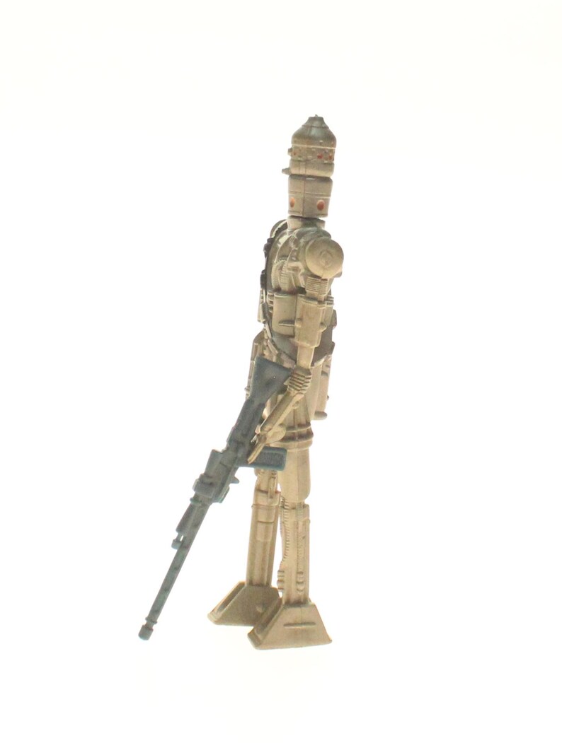 May include: A beige and gray plastic action figure of a Star Wars character, a droid, holding a blue and gray blaster rifle. The droid has a dome-shaped head with a red eye.