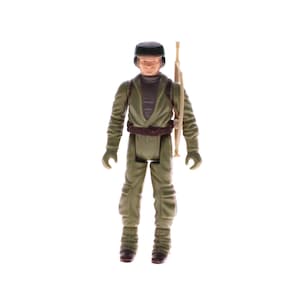 May include: A vintage action figure of a character in a green uniform with a brown belt and a black cap. The figure is holding a tan-colored rifle. The figure is standing on a white background.