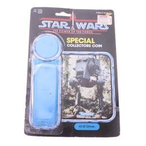 May include: A vintage Star Wars "The Power of the Force" collectible coin, packaged on a black and blue card. The card features an image of an AT-ST Driver and the Kenner logo. The coin is encased in a blue plastic bubble.