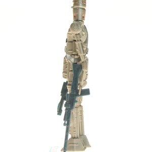 May include: A beige and gray action figure of a robot with a large head and a gun in its hand. The robot is standing on a white base.