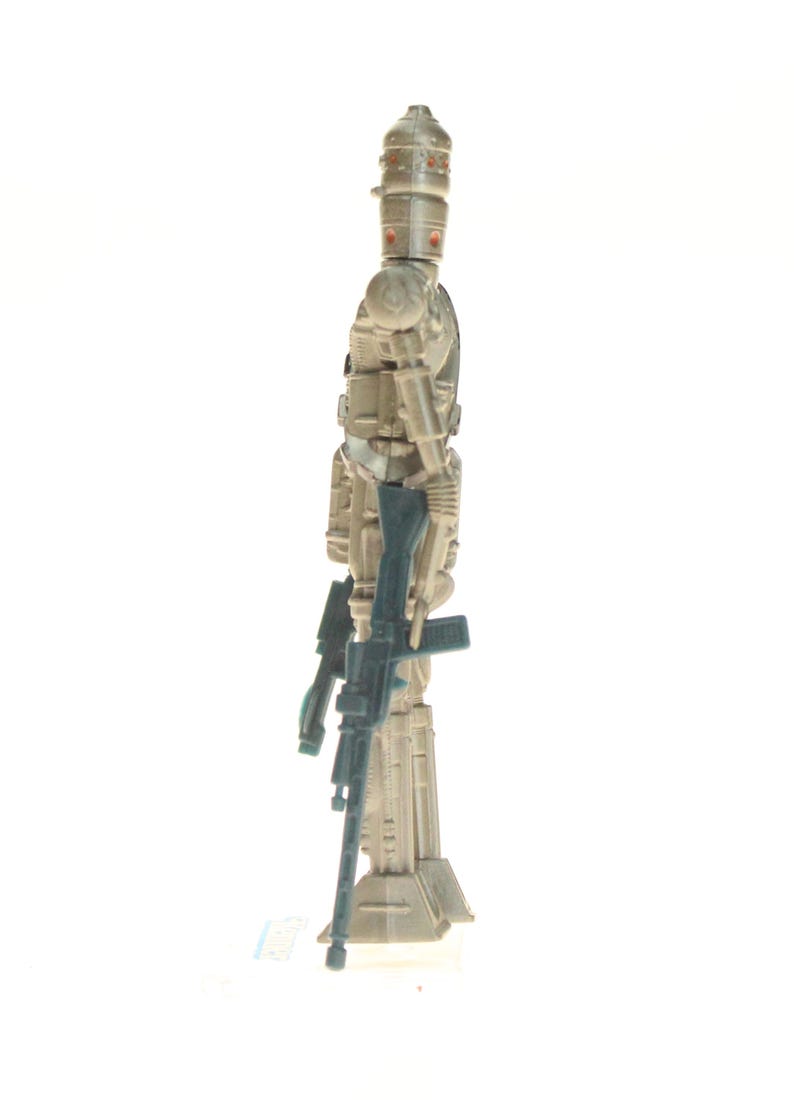 May include: A beige and gray action figure of a robot with a large head and a gun in its hand. The robot is standing on a white base.