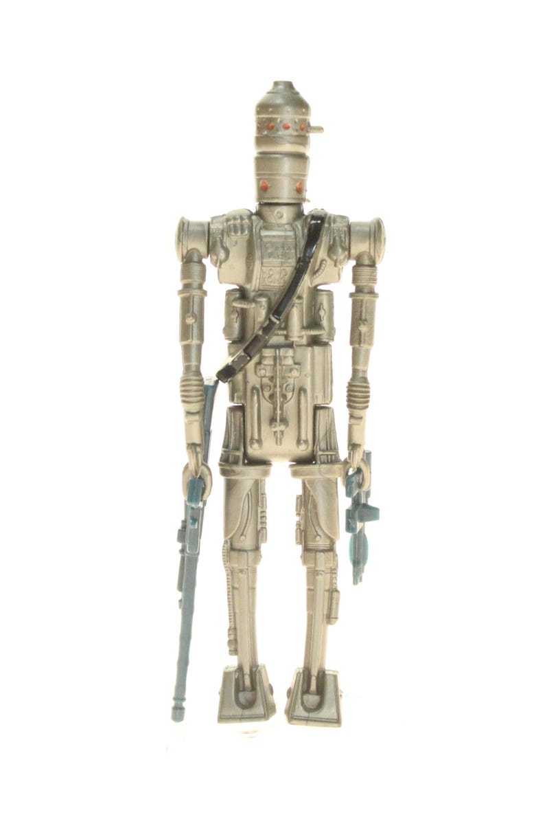 May include: A silver and gray action figure of a robot with a helmet and a blaster in each hand. The robot is standing on a white background.