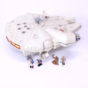 May include: A white plastic Millennium Falcon toy spaceship featuring a top-mounted grey laser cannon and a circular radar dish. Below the ship are six small character figurines, including a brown Wookiee, a gold droid, and four human figures.