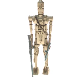 May include: A beige plastic action figure of a Star Wars character, a droid, holding two blue blasters. The droid has a dome-shaped head with a silver design.