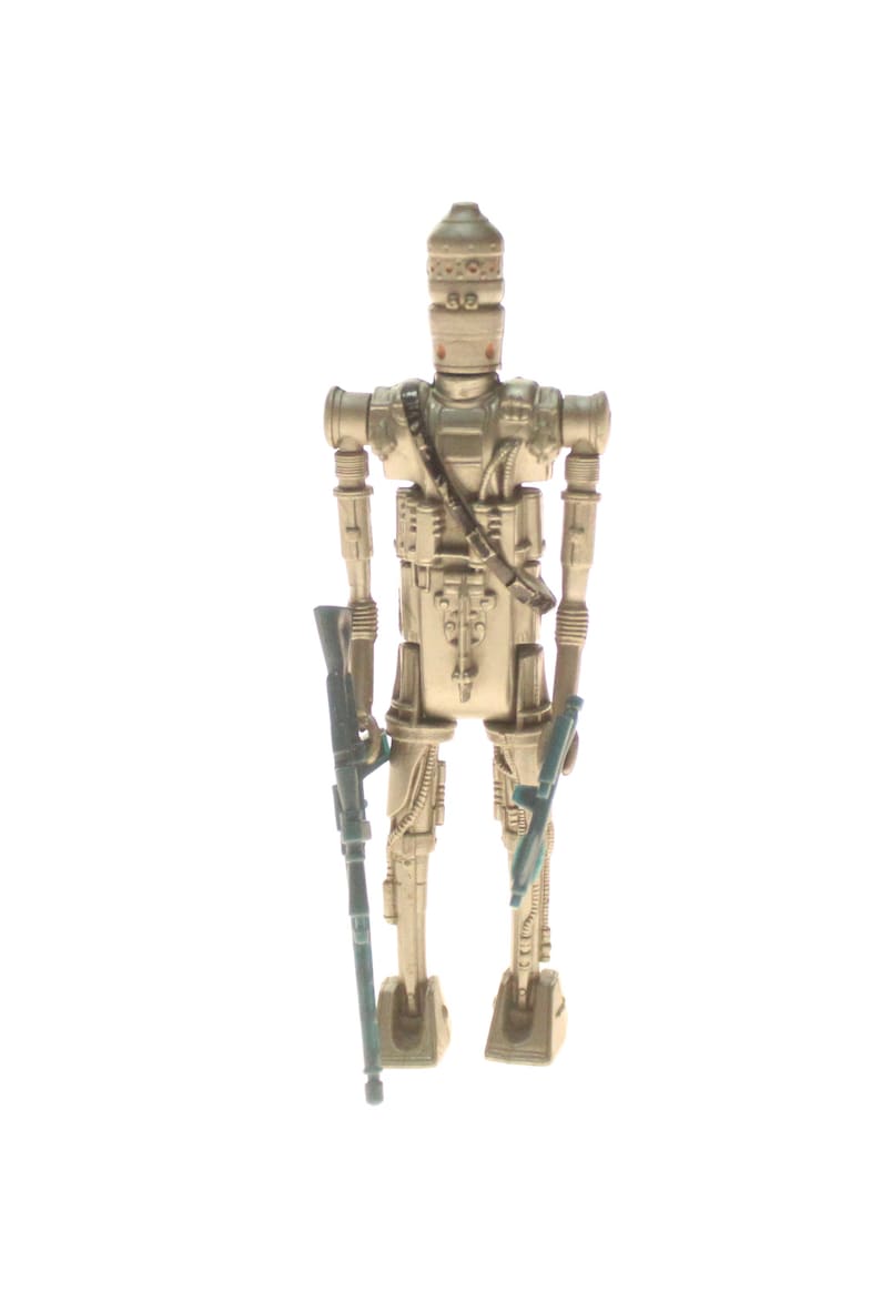 May include: A beige plastic action figure of a Star Wars character, a droid, holding two blue blasters. The droid has a dome-shaped head with a silver design.