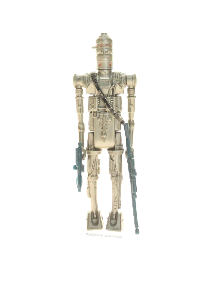 May include: A silver-colored action figure of a robot with a blue blaster in each hand. The robot has a helmet with red eyes and a black strap across its chest.