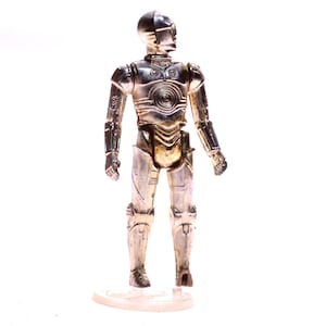 May include: A metallic gold and silver action figure of a humanoid robot. The figure has a detailed design with visible joints and a circular pattern on the chest. It stands on a clear plastic base, showcasing a classic sci-fi collectible.