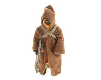 Könnte beinhalten: Braune Actionfigur eines Star Wars Tusken Raiders, der einen Kapuzenmantel und einen Bandolier trägt. Die Figur hält ein graues Gewehr. Die Figur steht vor einem weißen Hintergrund.
