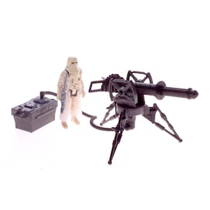 May include: A vintage action figure set comprising a black tripod-mounted cannon, a grey power supply, and a white and tan figure in a snowsuit. The cannon has a long barrel and detailed features.