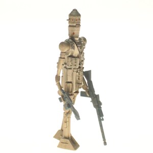 May include: A beige and brown plastic action figure of a Star Wars character, a droid, holding a blaster rifle in one hand and a smaller blaster in the other. The droid has a dome-shaped head with a visor and a body with multiple details.