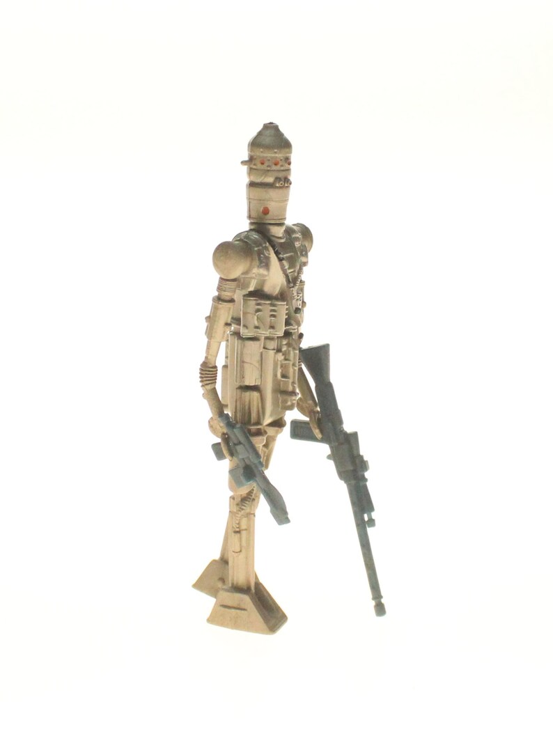 May include: A beige and brown plastic action figure of a Star Wars character, a droid, holding a blaster rifle in one hand and a smaller blaster in the other. The droid has a dome-shaped head with a visor and a body with multiple details.