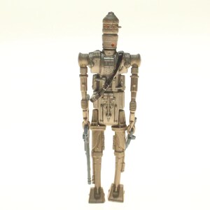 May include: A beige and silver action figure of a robot with a large head and a gun in each hand. The robot is standing on a white background.