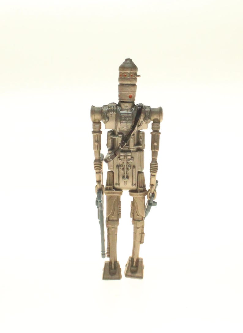 May include: A beige and silver action figure of a robot with a large head and a gun in each hand. The robot is standing on a white background.