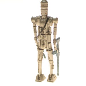 May include: A beige and silver action figure of a Star Wars character, a droid, holding a blue and gray blaster rifle. The droid has a tall, cylindrical head with a small, round face.